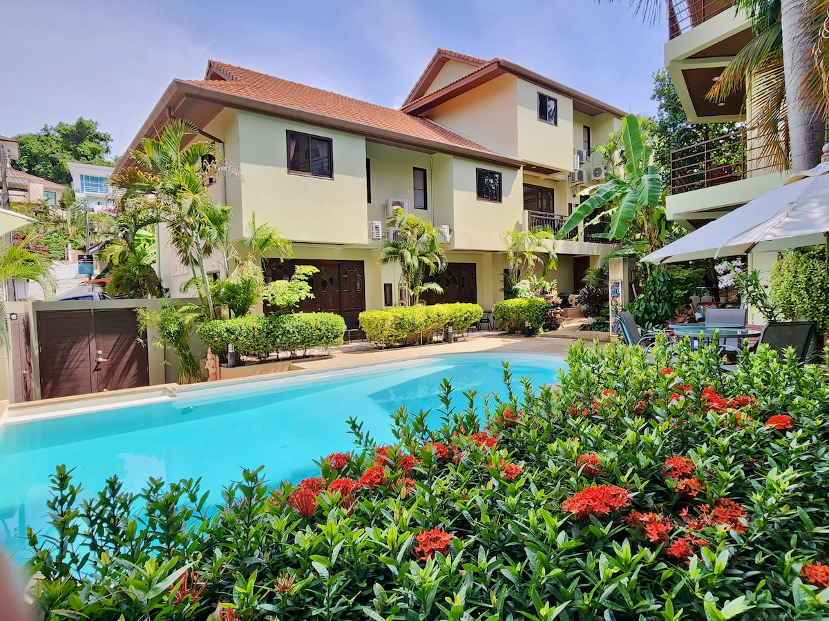 A serene view of the complex is presented, featuring lush greenery and colorful flowering plants bordering a clear swimming pool. The two-story buildings are surrounded by tropical palms and landscaping, creating a tranquil atmosphere, with shaded seating areas visible near the pool.