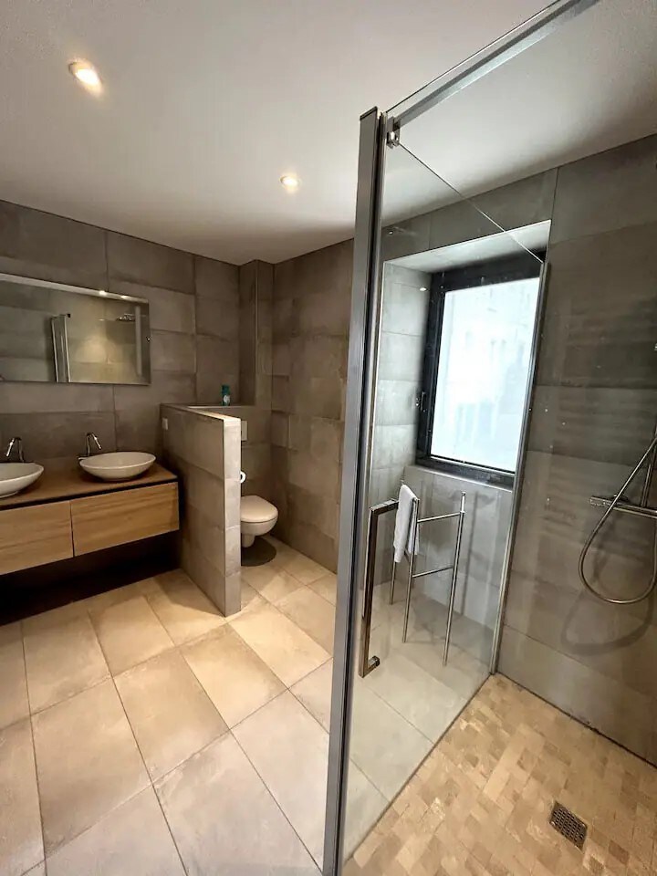 The bathroom features a spacious layout with a shower area separated by glass. Two modern sinks are mounted on a wooden vanity. Natural light enters through a window, and neutral-toned tiles cover the gray walls and floor.