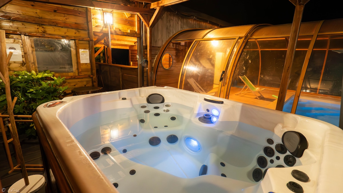 A well-lit outdoor jacuzzi is featured under a wooden pergola. Soft blue lighting adds a calming ambiance, while the surrounding area includes colorful plants and transparent panels that provide shelter and visibility to the heated pool area.