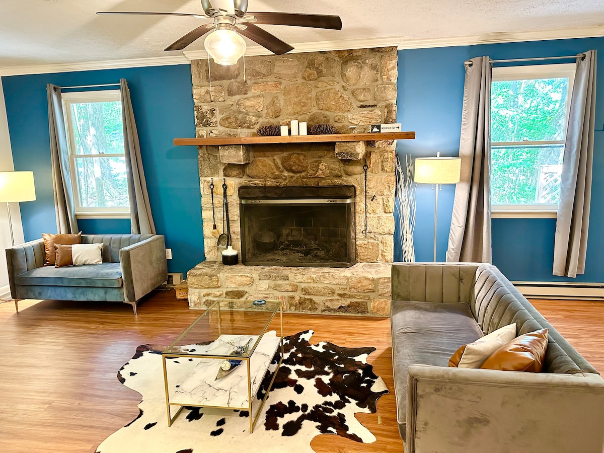 A spacious living area features a large stone fireplace as the focal point, surrounded by comfortable seating. Windows bring in natural light, while a coffee table rests on a patterned rug. Grey furnishings complement the blue wall, creating a cozy ambiance.