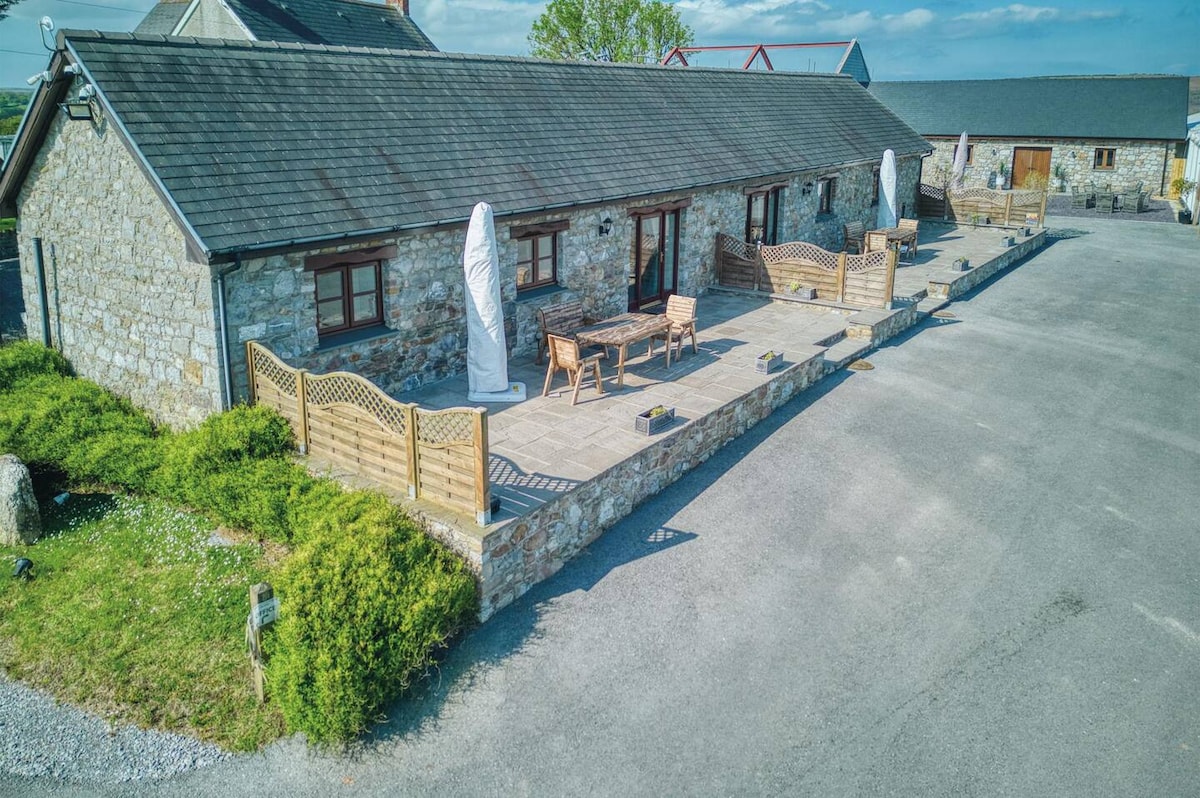 A stone exterior cottage is shown, featuring a spacious patio with wooden furniture. Surrounding greenery provides a natural border, while gravel pathways lead through the area. The structure's roof is dark, contrasting with the lighter stone walls, and the overall design reflects a rustic charm.