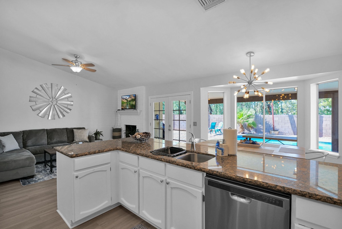 A modern kitchen space features a granite countertop with stainless-steel appliances. A comfortable seating area is visible, along with a light fixture above. Large windows provide natural light and views of the backyard pool area, connecting indoor and outdoor environments.