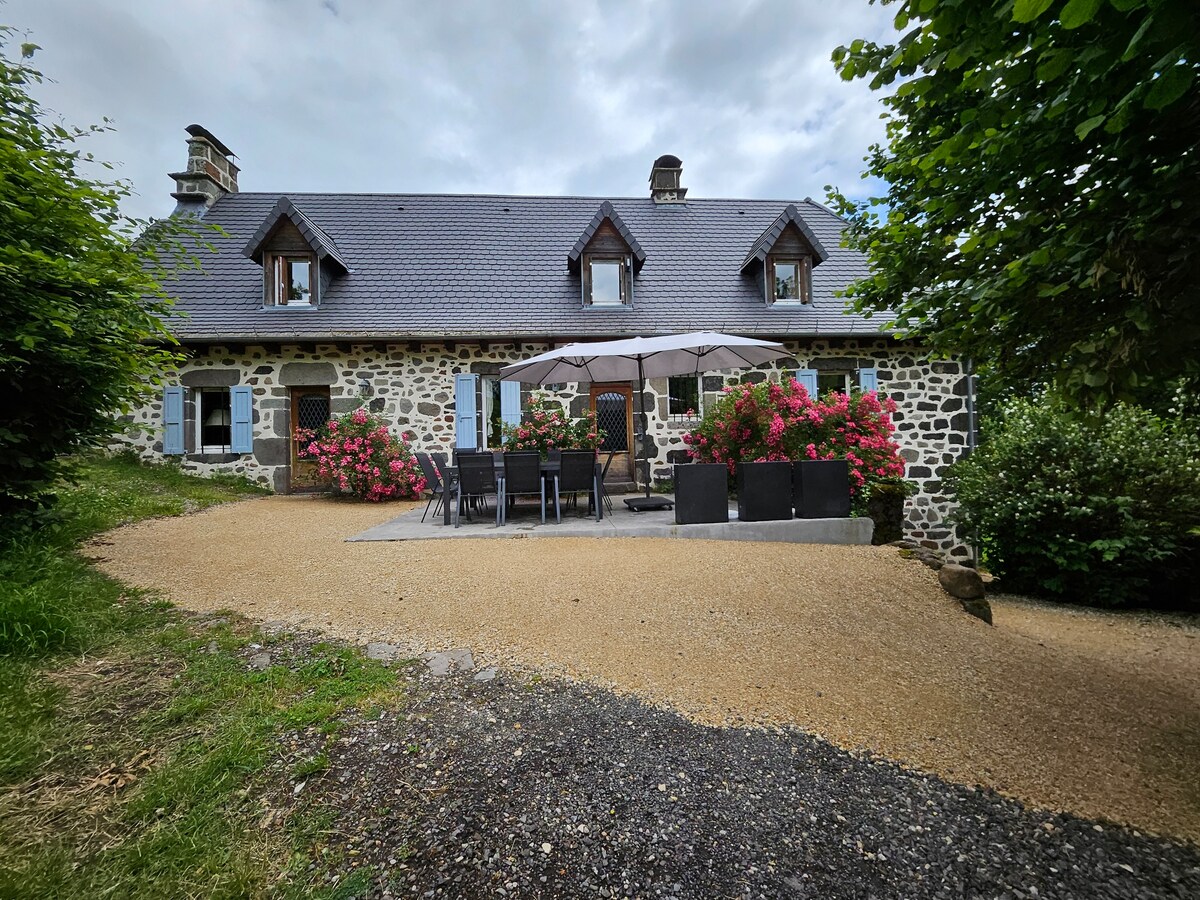 The exterior of a spacious stone house is framed by lush greenery and vibrant flowering plants. A large umbrella shades an outdoor dining area featuring a table and multiple chairs. A gravel pathway leads toward the entrance, surrounded by well-maintained gardens.