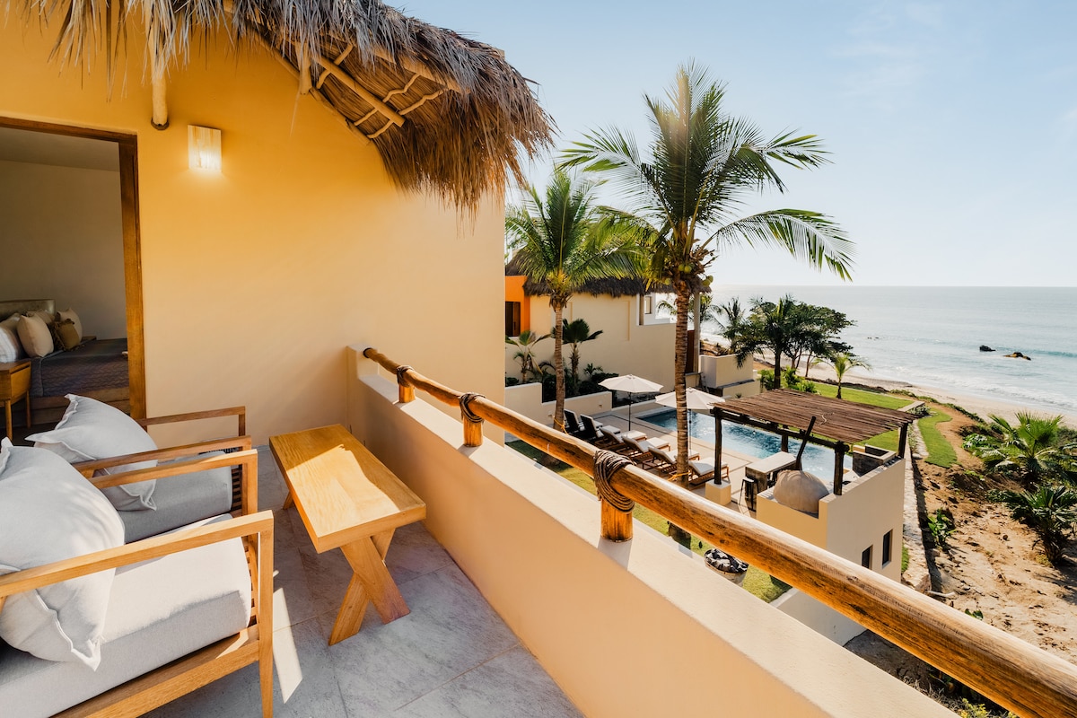 A balcony view features a wooden table and two armchairs, overlooking lush palm trees and a glistening ocean. The infinity pool and outdoor living space are visible, providing a serene setting with natural light illuminating the area.