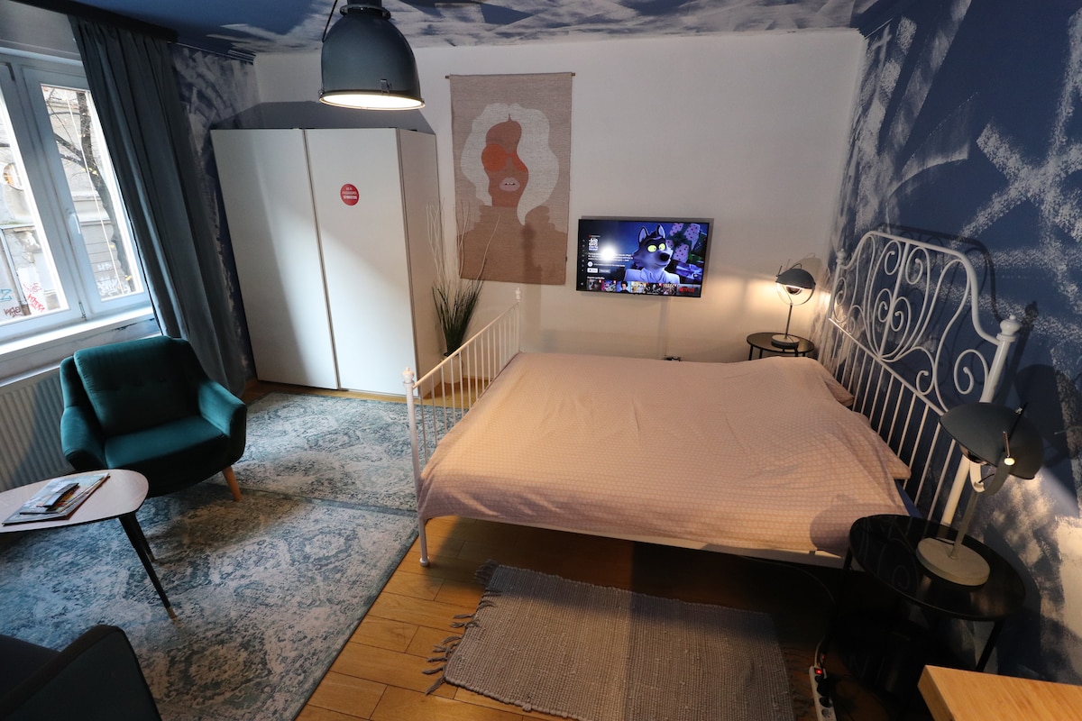 A studio apartment features a spacious bed with a soft beige coverlet, situated against a mural of blue clouds. A compact seating area includes a green armchair and a small table. A wall-mounted TV and a wardrobe are visible, enhancing the cozy ambiance.