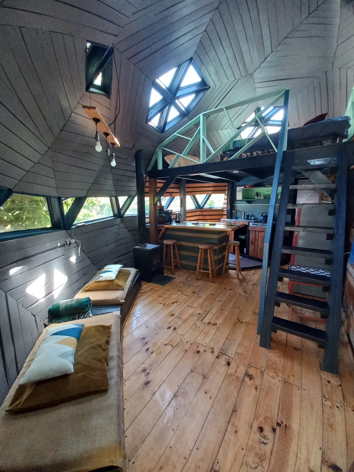The interior of the rustic dome showcases sloped wooden walls, complemented by large windows allowing natural light to fill the space. A cozy sitting area features cushions on a bench, while a ladder leads to a sleeping loft. The kitchenette is visible in the background.