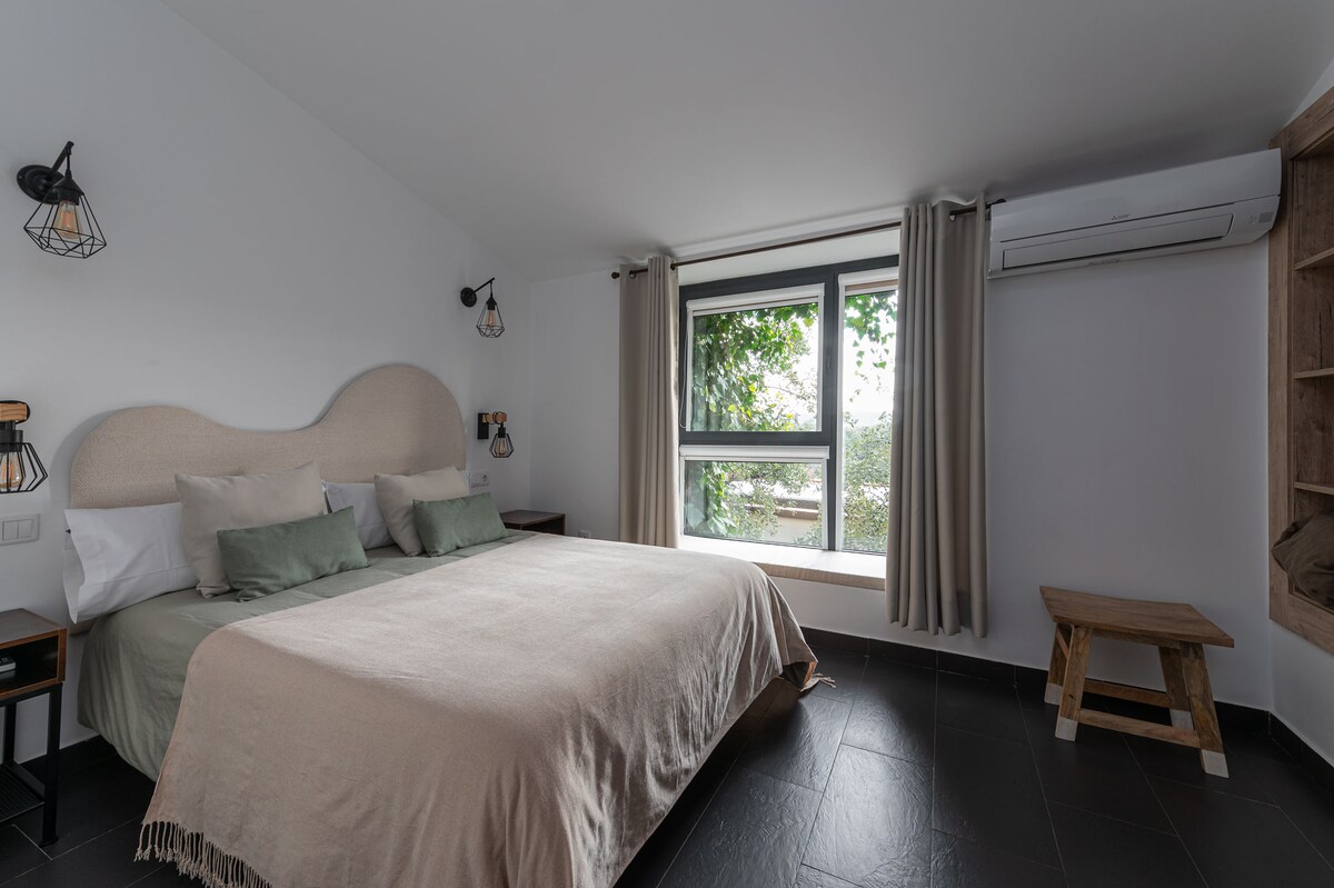 A spacious bedroom contains a large bed dressed in neutral linens, flanked by two bedside tables. Natural light enters through a large window, framed by light curtains. A comfortable seating area is created with a wooden bench, and air conditioning is positioned above the bed.
