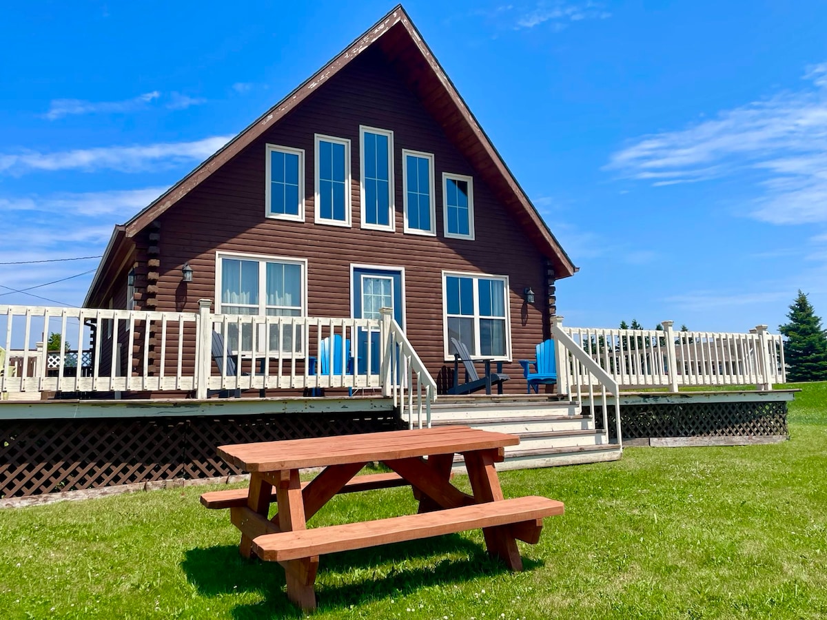 A cozy cedar log cabin is shown with a spacious deck featuring blue outdoor seating and views of the surrounding area. A picnic table is positioned on a grassy lawn, creating an inviting space for outdoor dining or relaxation.
