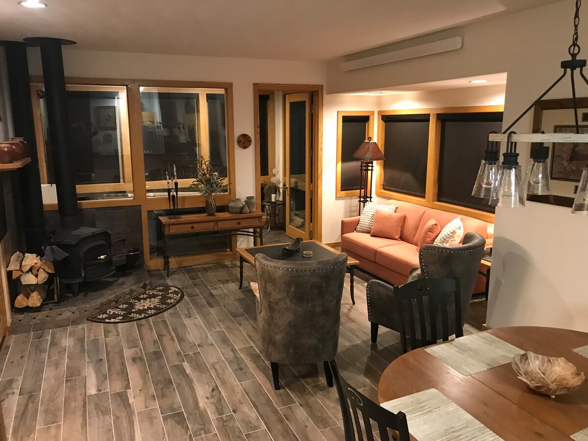 A cozy living area features a wood-burning stove and is furnished with a coral-colored sofa accented by pillows. Two plush chairs and a wooden dining table are visible, with large windows allowing natural light and views of the outdoors.