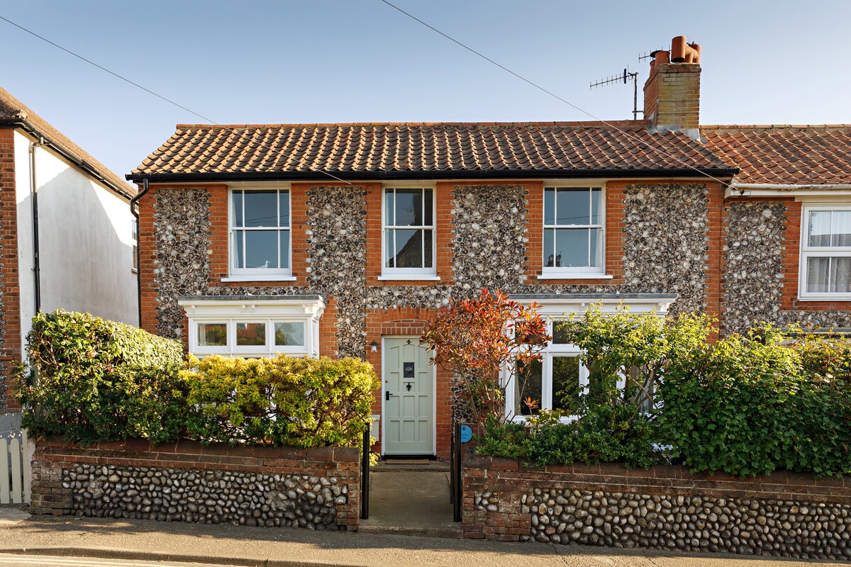 A charming cottage exterior is seen, featuring a stone and brick façade framed by lush greenery and shrubs. The entrance boasts a central door flanked by windows, offering a warm welcome. A neatly maintained pathway leads to the front entrance, enhancing the inviting appearance.