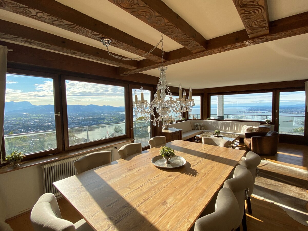 A spacious dining area features a large wooden table surrounded by eight comfortable chairs. A crystal chandelier hangs above, complementing the natural light that floods in through panoramic windows. Stunning views of the surrounding mountains and lake are visible, enhancing the elegant ambiance.