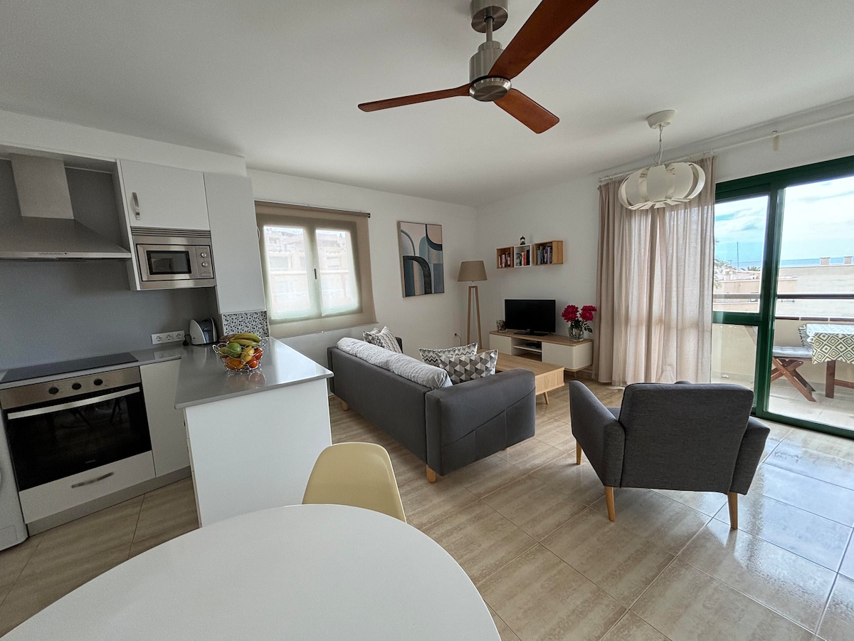 A modern living area features a comfortable sofa paired with an armchair, a coffee table, and a television. Natural light filters through large windows, illuminating the space. A dining table is positioned nearby, complementing the open layout of the room.
