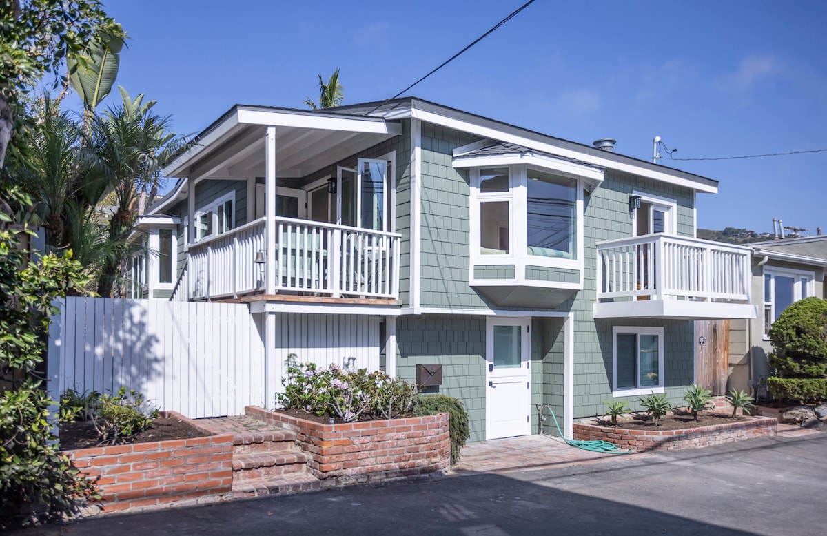 The exterior of a charming two-story cottage is seen, featuring a freshly painted green facade with white trim. Two balconies are positioned on the upper level, while a white door leads to the lower level. Lush landscaping surrounds the brick pathway and entrance.