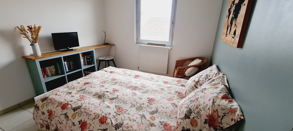 A serene bedroom features a spacious bed adorned with floral bedding. A small wooden desk stands against the wall, complemented by a television. A cozy chair is positioned in the corner, and a vase of dried flowers adds a touch of warmth to the space.