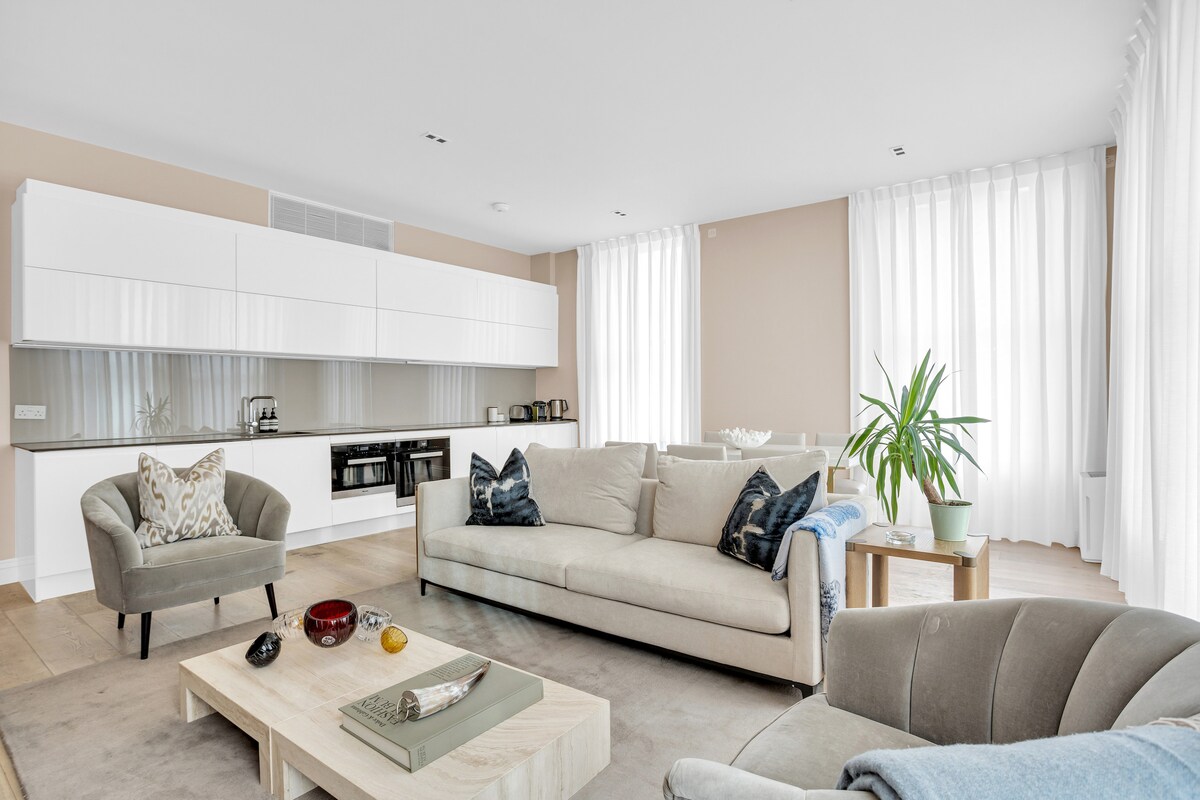A light-filled living area is furnished with a spacious sofa and an accent chair. A large coffee table sits in the center, decorated with a few small items. Floor-to-ceiling windows allow for natural light and are draped with sheer white curtains.