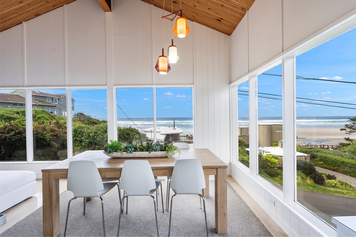 A dining area features a wooden table surrounded by six chairs, with natural light streaming through large windows. Scenic views of the ocean and nearby landscape are visible, complemented by a modern light fixture hanging from a vaulted ceiling.