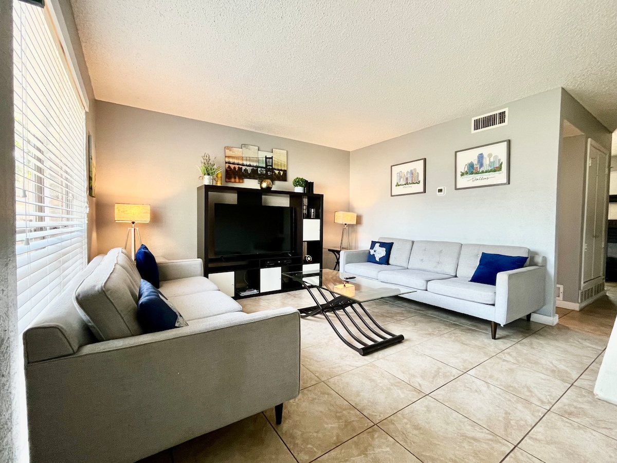 A light-filled living room features two modern sofas in soft gray, with blue accent pillows. A wall-mounted television is positioned on a black media console, complemented by decorative plants and artwork. Tile flooring adds a polished finish to the welcoming space.