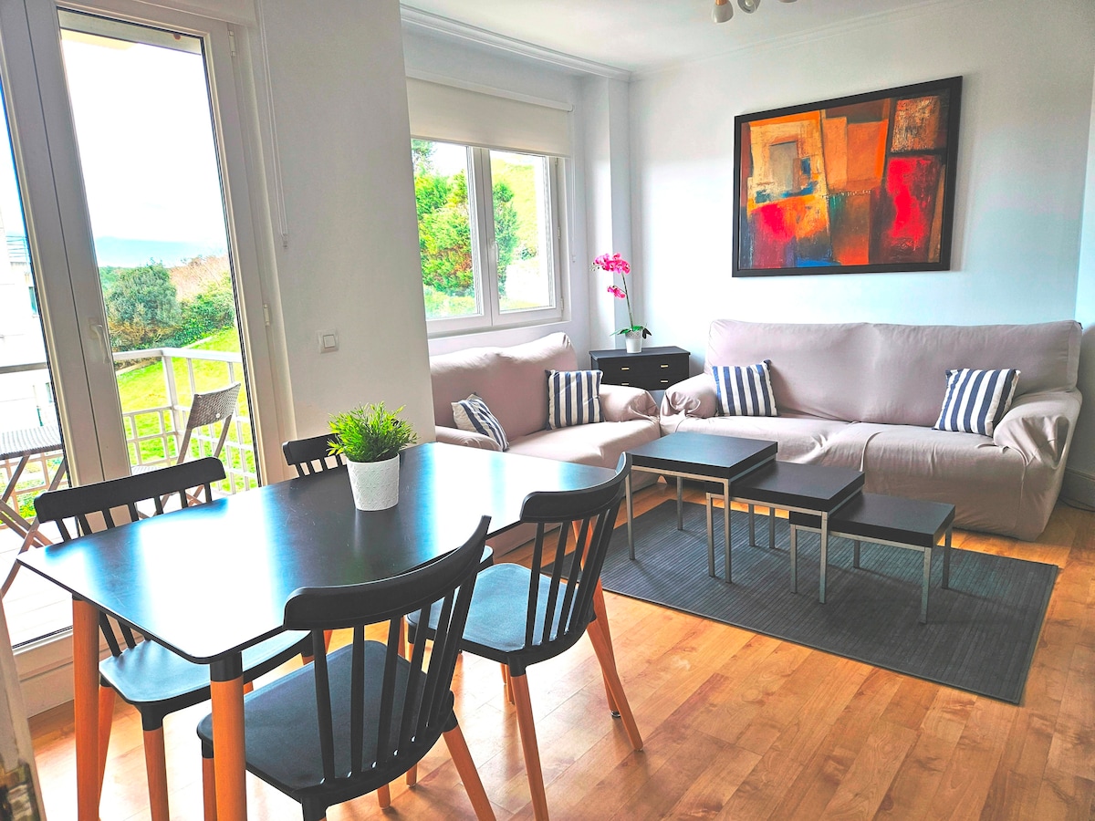 A bright living area features large windows allowing natural light to fill the space, showcasing a modern dining table with six black chairs. Two comfortable sofas, adorned with striped cushions, are positioned near a vibrant abstract painting on the wall, creating an inviting atmosphere.