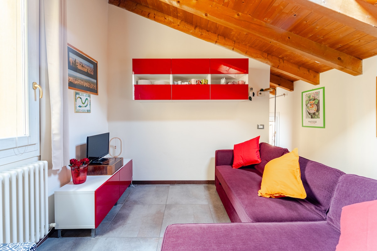 A cozy living area is characterized by a purple sectional sofa adorned with bright orange pillows. A sleek white television unit is positioned against a wall, while a vibrant red shelving unit adds a pop of color. Natural light filters in through the nearby window.