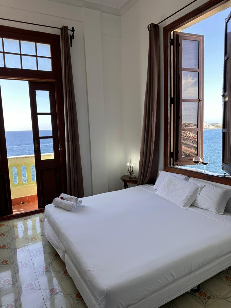 A bright room features a spacious bed dressed in white linens. Large windows with wooden shutters open to reveal an ocean view, allowing natural light to fill the space. Elegant candle holders stand on a nearby table, contributing to a serene ambiance.