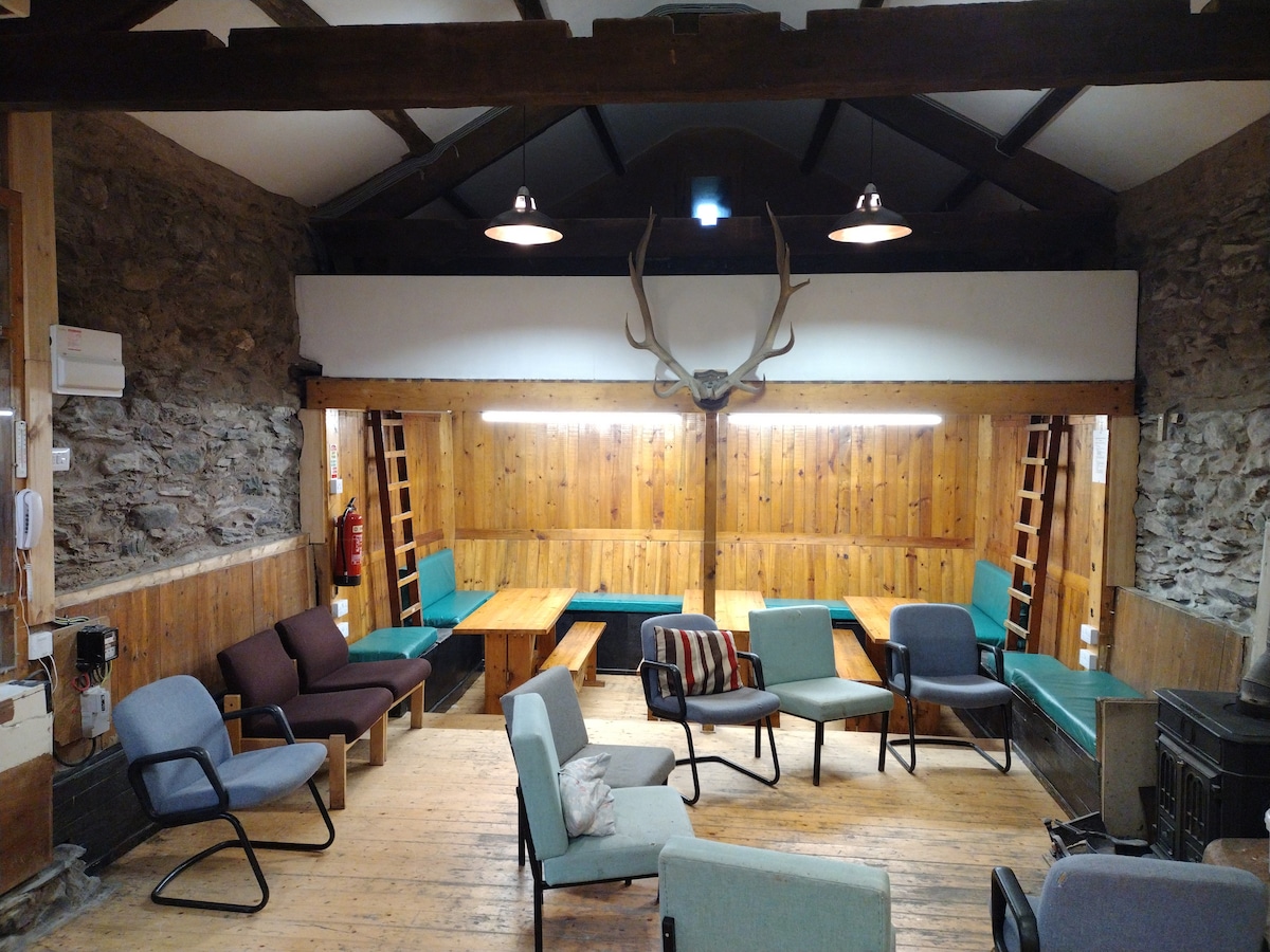 A communal lounge area is depicted with wooden walls and beam ceilings. Multiple comfortable chairs are arranged around rustic wooden tables. A prominent deer antler mount decorates the wall, while soft lighting creates a cozy atmosphere. A wood-burning stove is visible, enhancing the rustic setting.