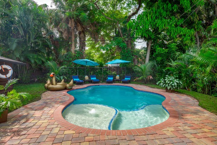 The outdoor area features a lagoon-shaped saltwater pool surrounded by lush greenery. Multiple lounge chairs are placed under large umbrellas, providing shaded seating. A playful inflatable floats in the pool, inviting relaxation and fun. The brick patio offers a warm and welcoming atmosphere.