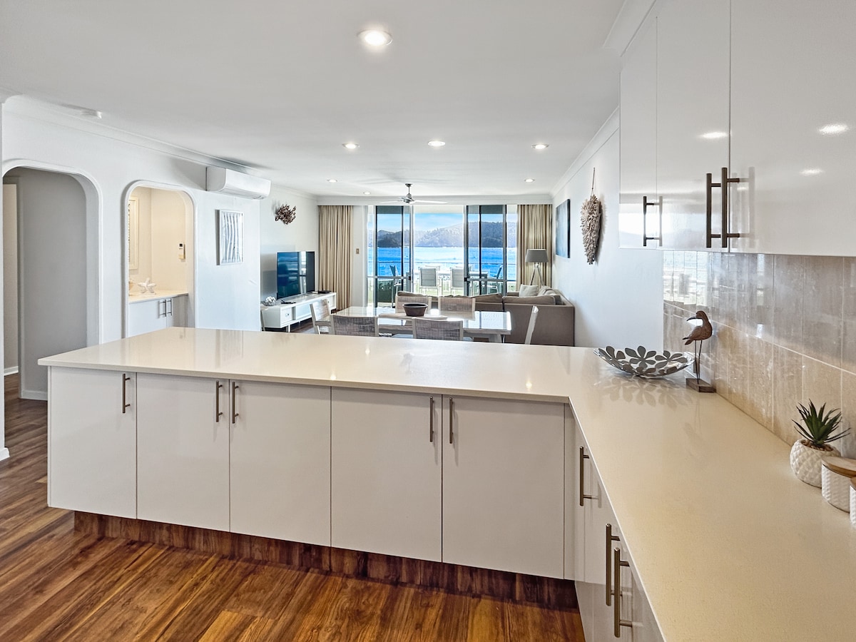 An open-plan kitchen area featuring modern cabinetry and a spacious countertop. Natural light filters through large windows, revealing a view of the ocean and the living area, which is furnished with a cozy couch and TV. The warm wooden flooring adds a welcoming touch.