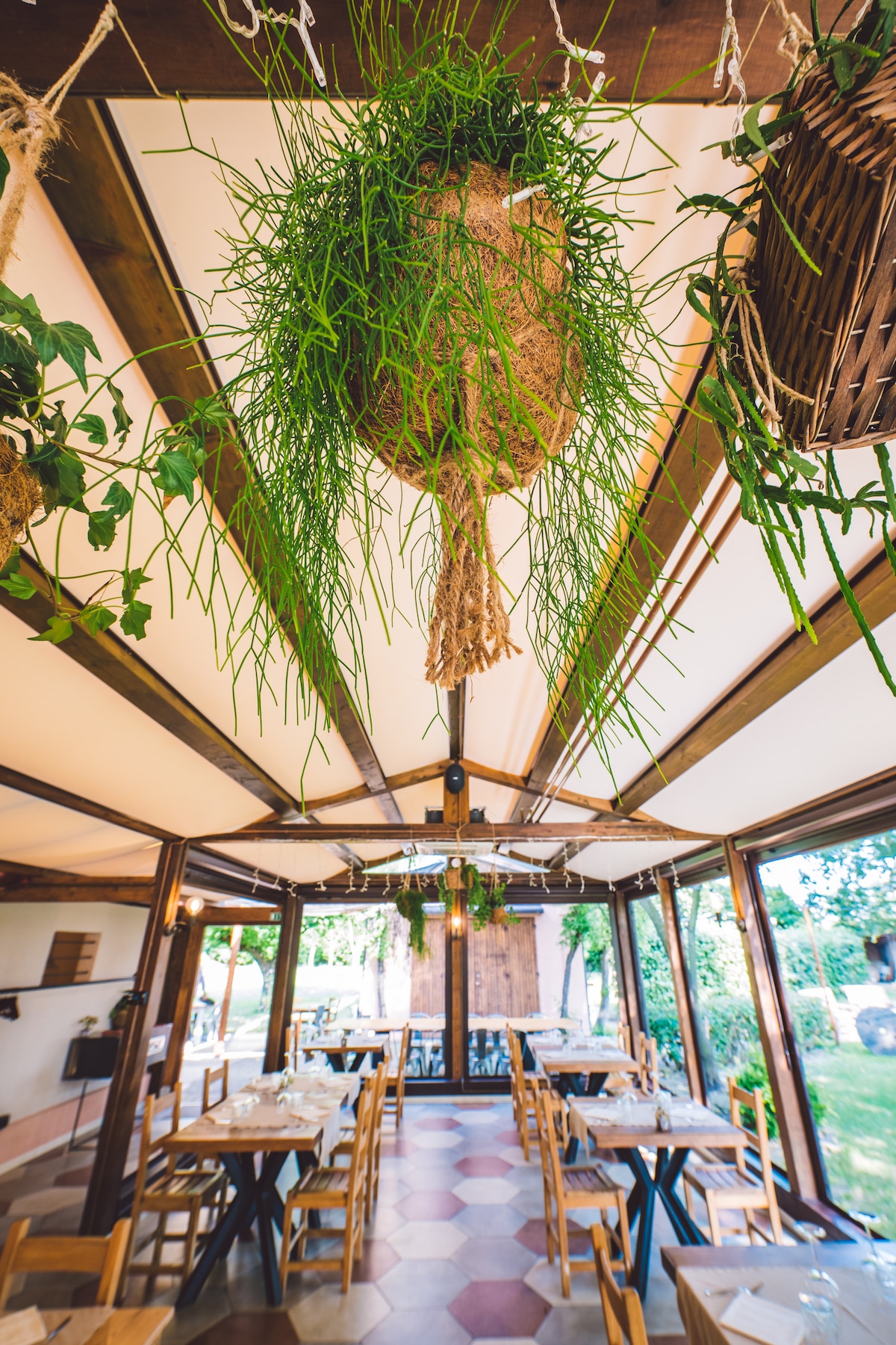 The dining area is characterized by wooden beams and large glass windows that create an open and airy environment. Hanging planters filled with greenery add a touch of nature, while rustic tables and chairs provide a welcoming space for dining.