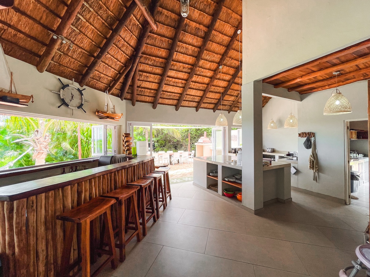 A spacious kitchen features a high thatched roof and large windows that allow natural light. Wooden bar stools are arranged at a rustic bar, and modern appliances are visible in the kitchen area. The layout accommodates interaction while cooking and dining.