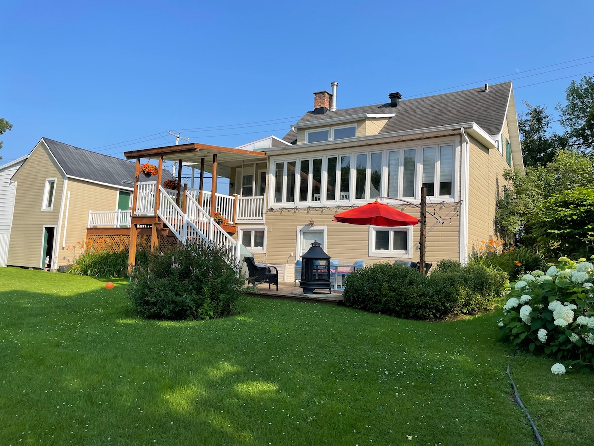 The exterior of the cottage is shown, featuring a two-story structure with large windows. A deck with a red umbrella offers outdoor seating. Lush green grass surrounds the home, complemented by well-maintained shrubbery and patio furniture. A neighboring building is visible in the background.