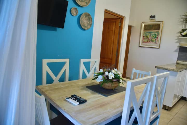 במרכז החדר יש שולחן אוכל גדול מעץ בהיר, מוקף בכיסאות לבנים עם ריפוד תואם לקירות, שיוצרים רצף כרומטי הרמוני.