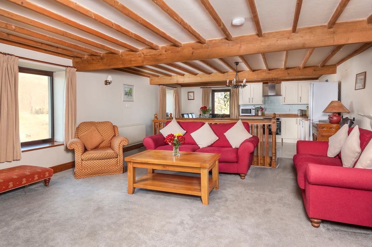 A cozy lounge area features polished wood beams overhead and is furnished with a pair of red sofas and matching armchair. A wooden coffee table sits center stage, while a bouquet of flowers adds a touch of color. Natural light pours in from a large window.