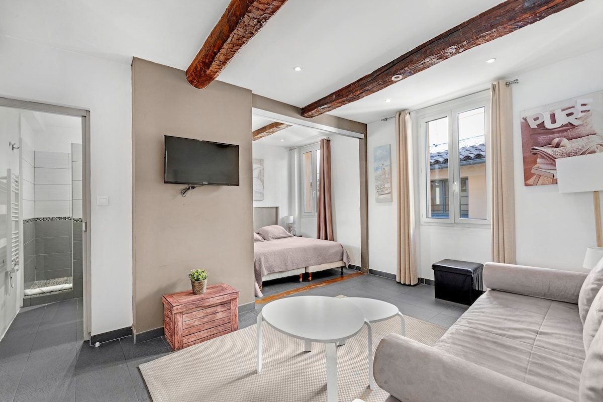 A bright and contemporary living space features exposed wooden beams and a soft color palette. A cozy seating area includes a light-colored sofa and a small round table. A wall-mounted television and framed artwork complement the decor, while the sleeping area is visible beyond a partition.