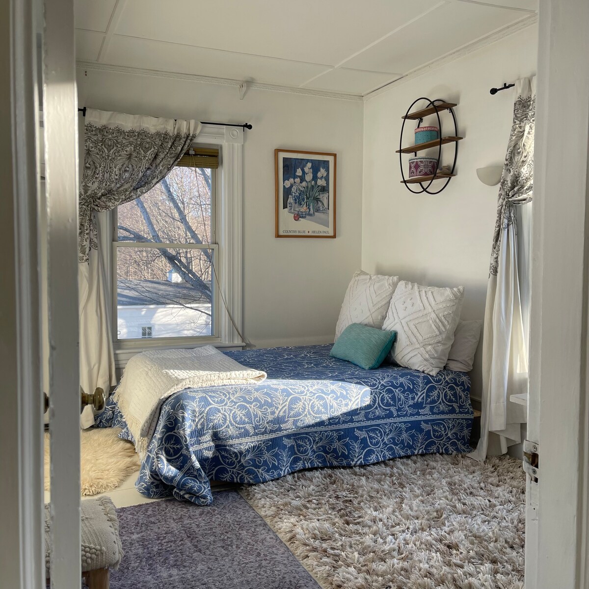 A room is presented with a cozy bed draped in blue and white textiles. Natural light streams through a window adorned with curtains, casting a soft glow. Decorative items, including wall art and a shelf, enhance the inviting simplicity of the space.