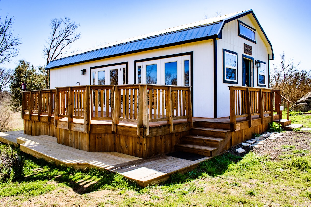 The exterior of the cabin features a welcoming wraparound deck made of wood, with a set of stairs leading to the entrance. Large windows allow natural light to fill the interior, and the building is surrounded by open green spaces and trees.