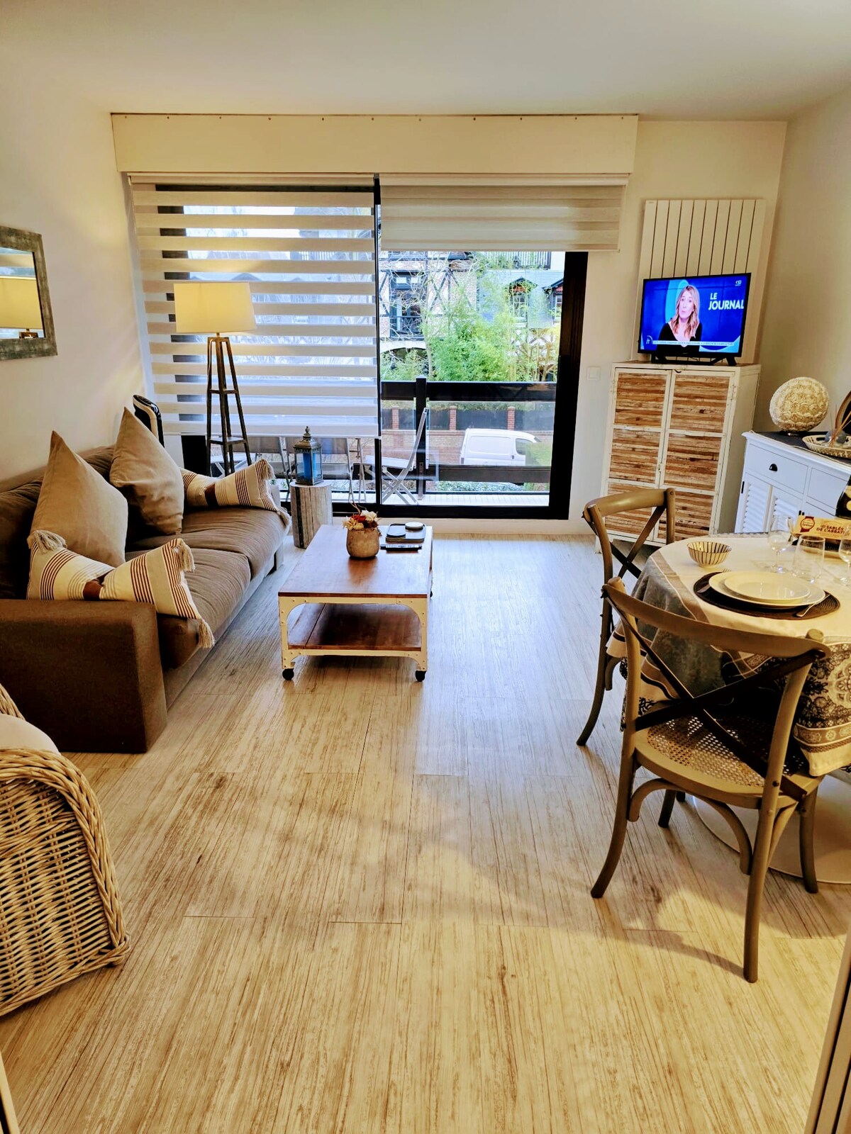 The living area is designed with a comfortable sofa and a decorative coffee table, complemented by natural light filtering in through large windows with horizontal blinds. A television is visible, along with a dining table set for four, enhancing the cozy atmosphere.