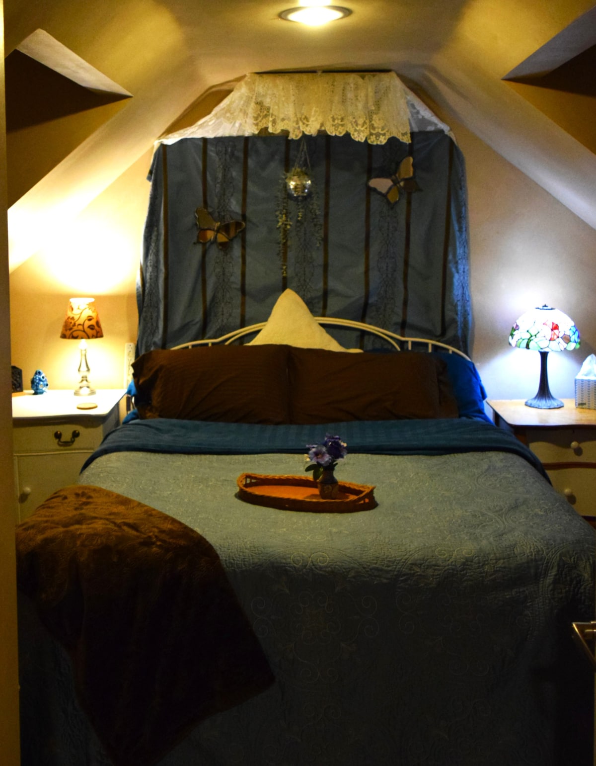 A cozy sleeping area is highlighted by a bed dressed in a blue quilt, accompanied by decorative pillows. Soft lighting from bedside lamps creates a warm ambiance, while a small tray holds fresh flowers on the nightstand, adding a touch of color and serenity.