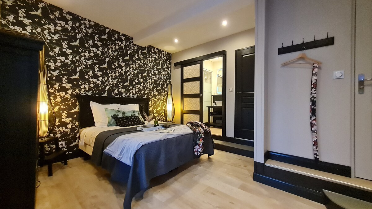A bedroom features a large bed adorned with contrasting linens. One wall is decorated with a floral wallpaper design, while the room is illuminated by softly glowing lamps. A glimpse of an ensuite bathroom can be seen through a partially open door.