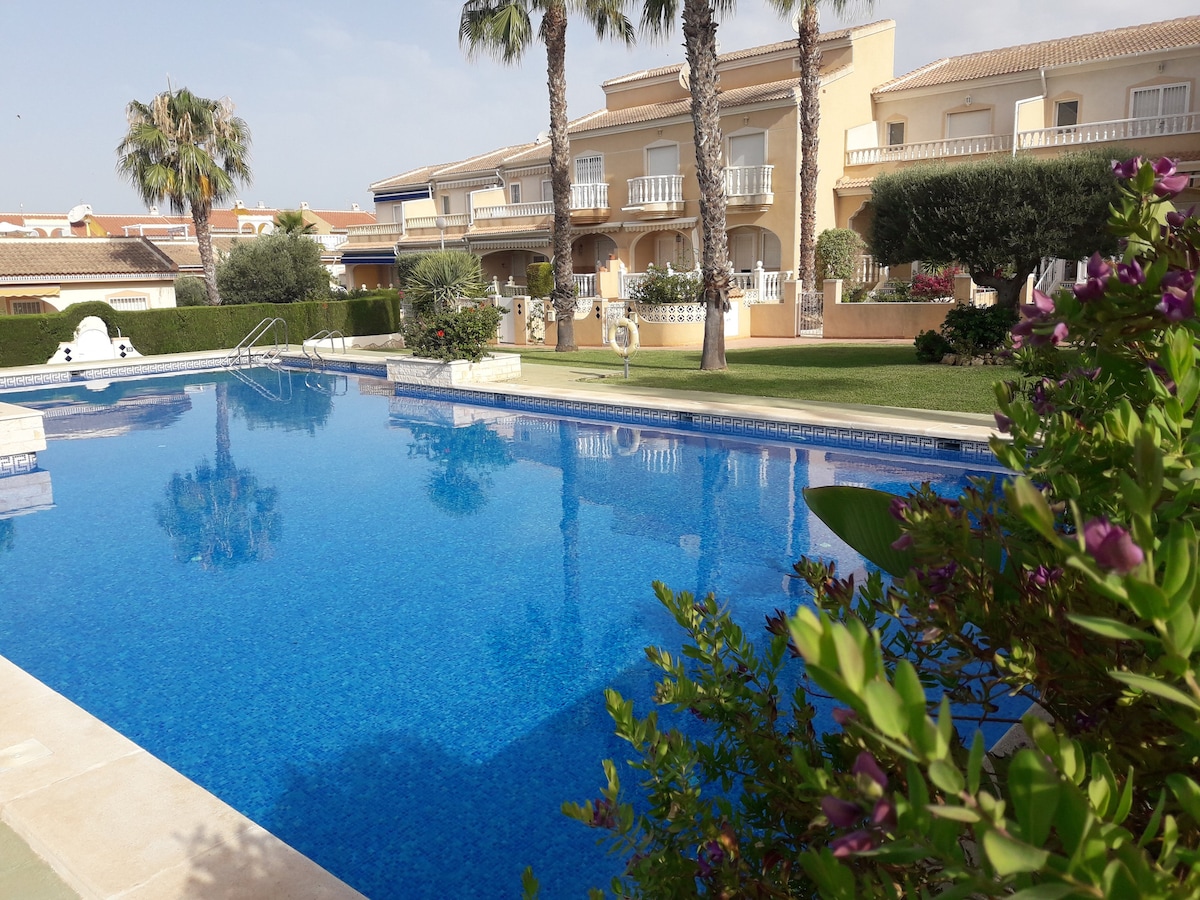 A large community swimming pool reflects the clear blue sky, surrounded by palm trees and well-maintained gardens. The pool area features loungers positioned along the edge, offering plenty of space for relaxation. Nearby buildings are visible, showcasing a residential setting.