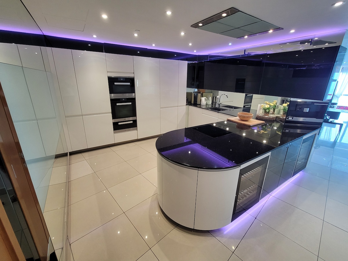 The modern kitchen features sleek cabinetry in high-gloss white and black. A large central island with a dark countertop is complemented by built-in appliances and ambient lighting along the edges. The polished tile flooring reflects the clean lines and contemporary design.