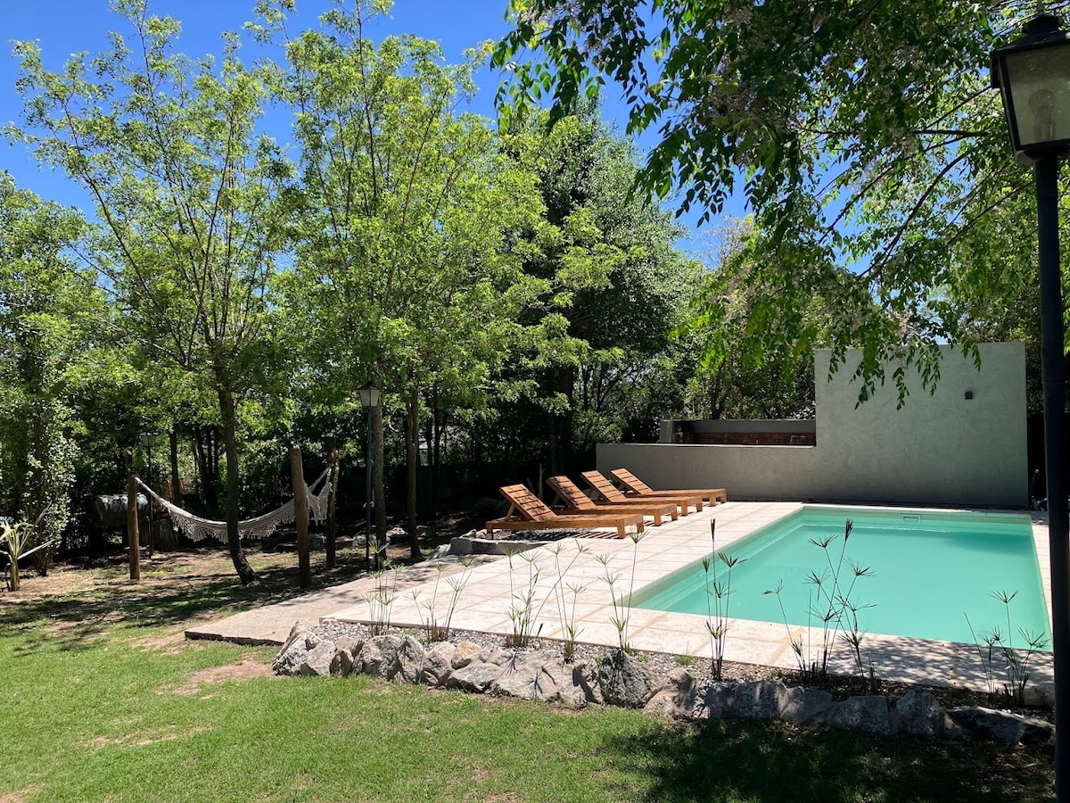 A serene garden area highlights a swimming pool surrounded by wooden lounge chairs. Lush green trees provide shade, while a hammock is visible nearby, inviting relaxation. The pool area features natural stone edging, and the sunlit scene showcases a calm, inviting outdoor space.