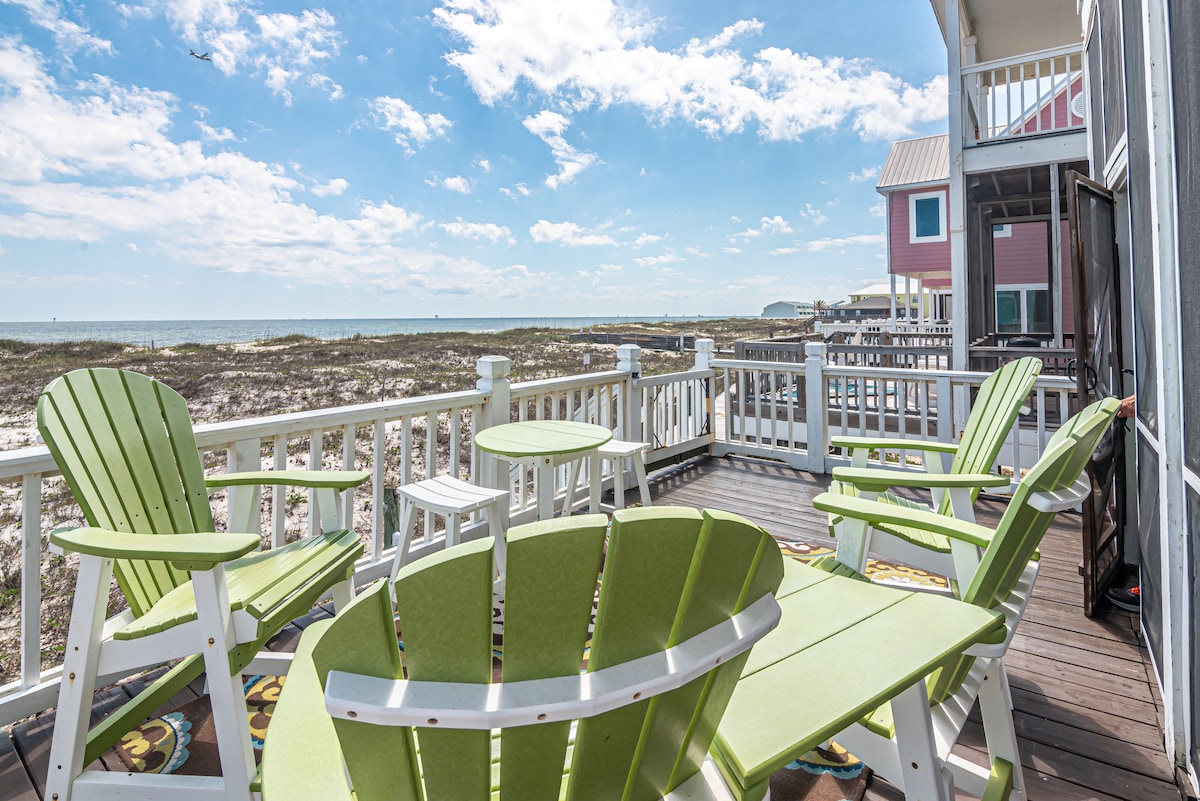A spacious deck overlooks a sandy beachfront, featuring light green outdoor furniture, including four chairs and a small round table. The scene captures expansive views of the coastline under a bright, partly cloudy sky, inviting relaxation in a serene setting.