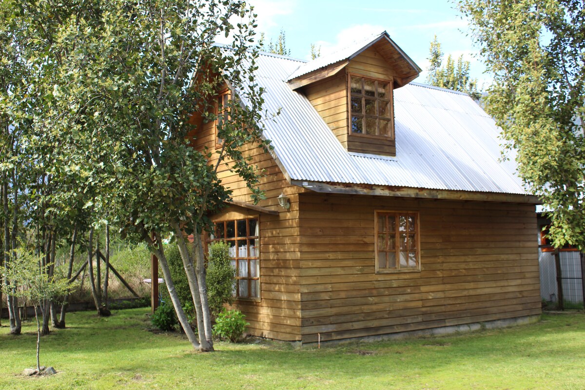 A wooden cabin is surrounded by trees and a grassy area. The structure features a gabled roof with a small upper window and multiple large windows that allow natural light to enter. The exterior showcases a rustic charm typical of natural settings.