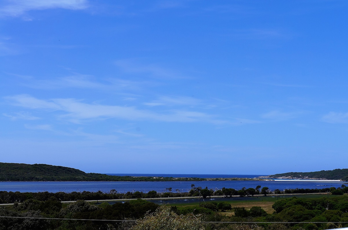 A panoramic view captures the serene waters of Wilson Inlet, edged by lush green landscapes and sandy shores. The sky appears vast and blue, creating a peaceful backdrop against the natural beauty of the coastal scenery.