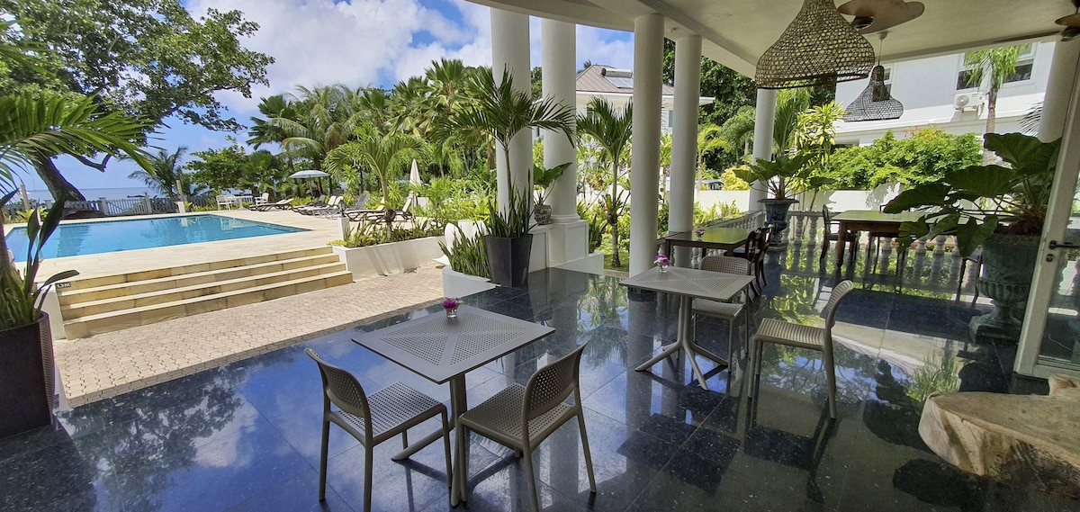 A covered outdoor area features two tables with chairs, set on polished stone flooring. Lush tropical plants surround the space, providing a natural backdrop. In the distance, a clear blue pool is visible, complemented by bright blue skies and scattered clouds.