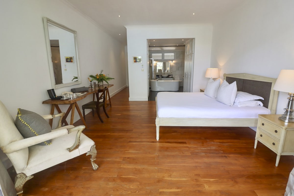 A spacious suite features a king-size bed with crisp white bedding, complemented by a wooden nightstand and a stylish lamp. A vintage mirror and a small dining table with chairs are present, while natural light fills the room through a nearby window, highlighting the warm wooden floors.