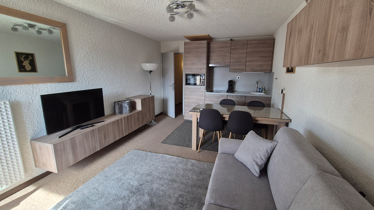 A cozy living area is presented with a grey sofa facing a modern TV unit. A glass dining table with black chairs is positioned adjacent to a compact kitchen featuring cabinetry and essential appliances. A soft area rug complements the warm tones of the decor.