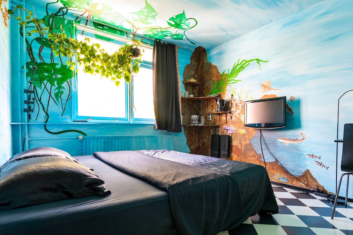 A bedroom is illuminated by natural light filtering through large windows, adorned with green plants and aquatic-inspired murals. A spacious bed with dark bedding is centrally placed, complemented by a television mounted on the wall and a small shelf displaying decorative items.