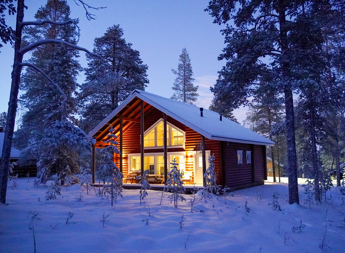 The log villa is enveloped in a serene winter landscape, with snow-covered ground and tall trees lining the property. Warm yellow light spills from the large windows, creating a welcoming glow against the evening sky.