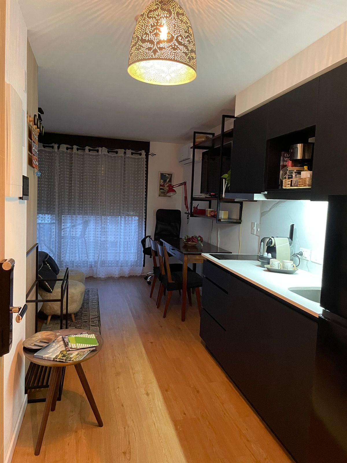 The kitchen area features modern black cabinetry and countertop, with essential appliances such as a kettle. A dining space with a wooden table and chairs is visible, alongside a cozy seating arrangement. Natural light filters through sheer curtains, enhancing the warm ambiance.